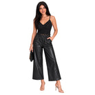 A woman modeling a black two-piece outfit consisting of a lace camisole top and high-waisted black faux-leather wide-leg pants, paired with nude high heels and holding a small black clutch, set against a plain white background.