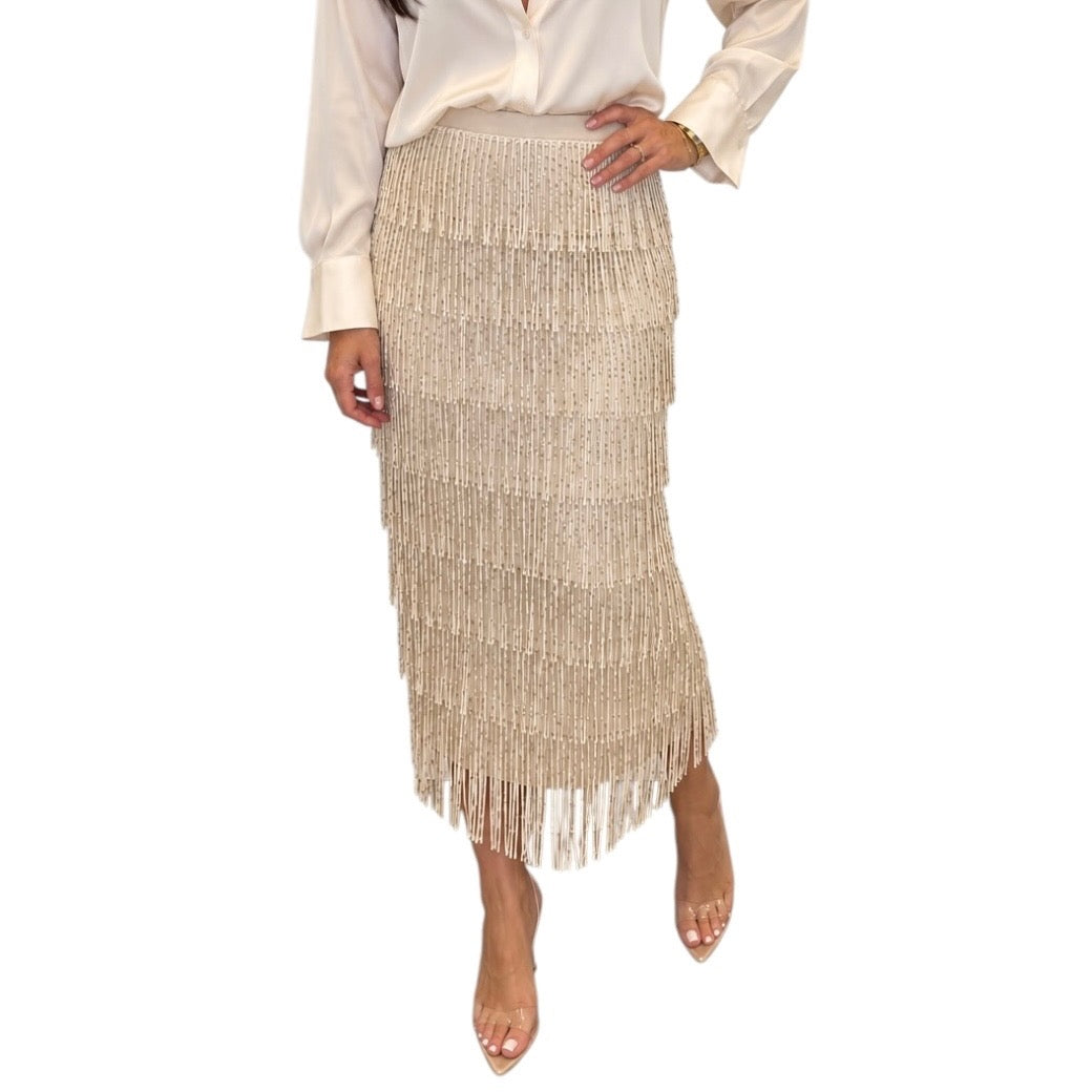 A model wearing a champagne beige fringed maxi skirt with a cream satin blouse and clear high-heeled sandals, shown against a plain white background.