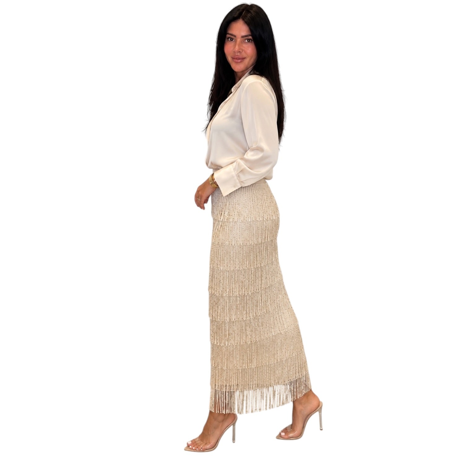 A woman modeling a long, shimmering champagne-gold fringe maxi skirt paired with a cream satin blouse and clear high-heeled sandals, shown against a solid white background.