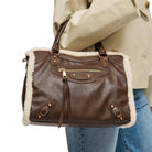 A brown faux-leather handbag with white shearling lining, carried on the shoulder of a person wearing a beige trench coat and blue jeans against a white background.