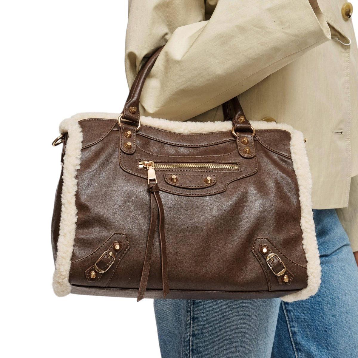 A brown faux-leather handbag with white shearling lining, carried on the shoulder of a person wearing a beige trench coat and blue jeans against a white background.