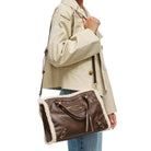 A person wearing a beige jacket and blue jeans carries a brown leather shoulder bag with white shearling lining and gold-tone hardware against a plain white background.