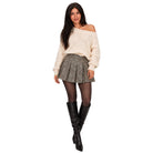 A woman modeling an off-the-shoulder cream knit sweater, a gray pleated mini skirt, sheer black tights, and black knee-high boots against a plain white background.