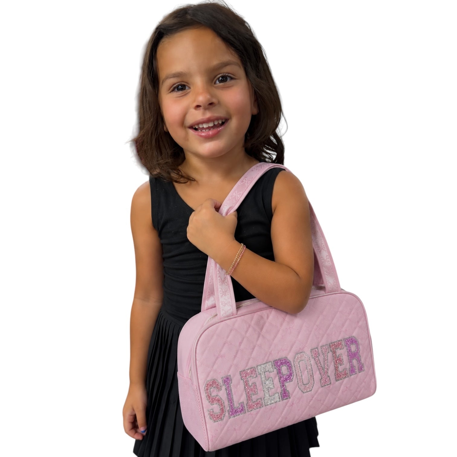 A young girl with dark hair smiling and holding a light pink quilted duffel bag with the word 'SLEEP OVER' in glittery letters, wearing a black sleeveless dress against a plain white background.