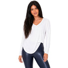 A woman modeling a white long-sleeve V-neck top and shiny dark navy leggings with white sneakers, standing against a plain white background.