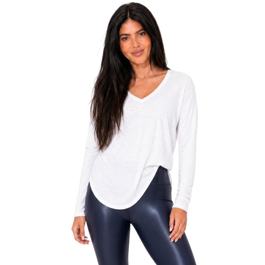 A woman modeling a white long-sleeve V-neck top and shiny dark navy leggings with white sneakers, standing against a plain white background.
