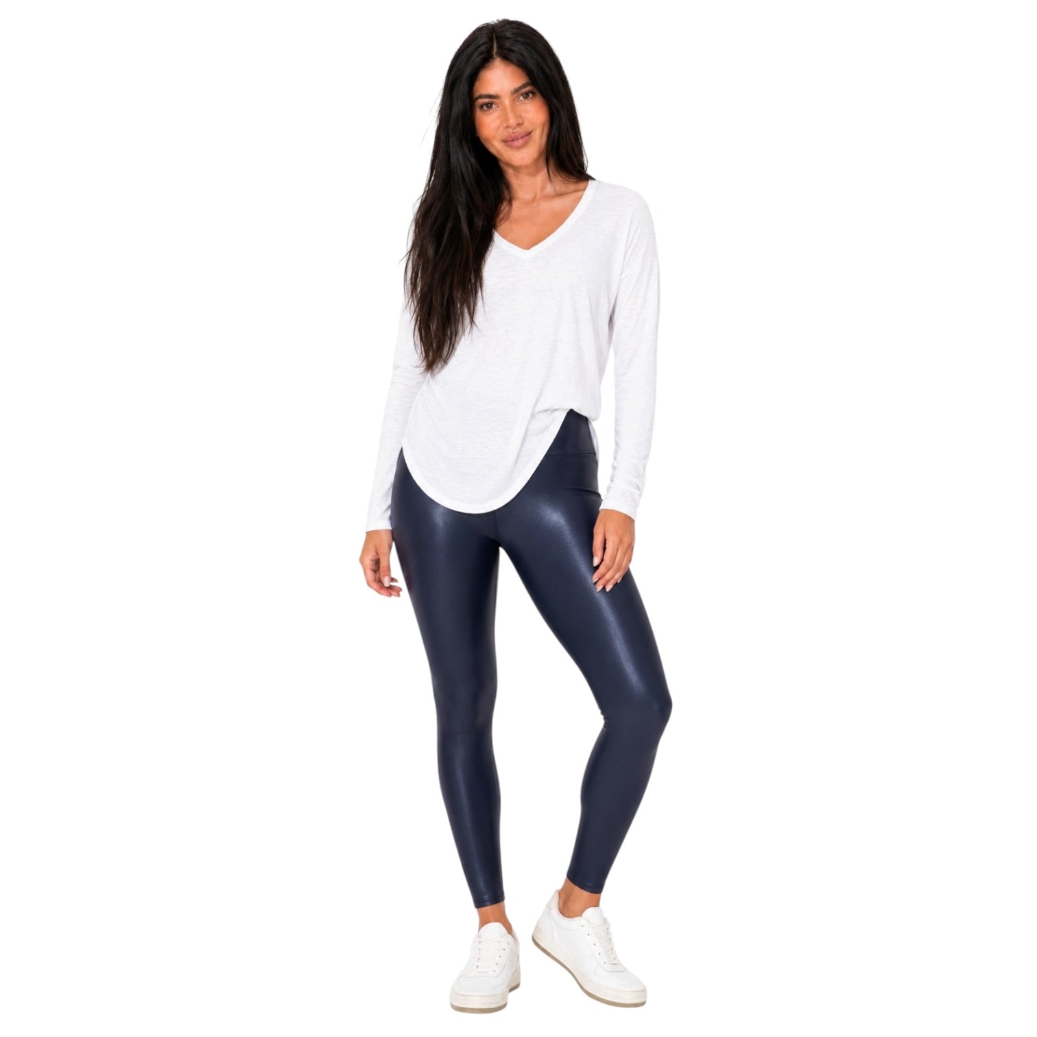 A woman modeling a white long-sleeve V-neck top and shiny dark navy leggings with white sneakers, standing against a plain white background.
