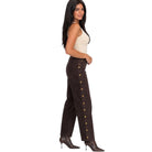 A woman modeling a pair of dark brown wide-leg pants with gold-tone decorative buttons down the side, paired with a cream tank top and brown pointed-toe heeled boots, set against a plain white background.