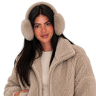 A woman wearing fluffy faux-fur earmuffs in a light beige color, paired with a matching beige sherpa fleece jacket and a cream ribbed sweater, set against a solid white background.
