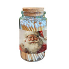A clear glass jar with a cork lid, filled with wooden matches topped with white tips, and wrapped with a printed label showing a festive Santa Claus portrait. The jar is centered on a plain white background.