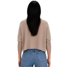 A woman viewed from behind wearing a cropped, light beige knit sweater with three-quarter sleeves and blue jeans against a plain white background.