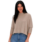 A woman modeling a light taupe knit mock-neck sweater with three-quarter ribbed sleeves, paired with blue jeans, against a solid white background.
