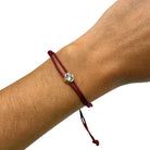 A close-up of a person's wrist wearing a dark red braided cord bracelet with a small round gold bezel-set clear stone.