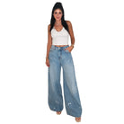 A woman modeling a pair of wide-leg light blue denim jeans with white paint splatter details, paired with a white crop top and gold sandals, set against a plain white background.