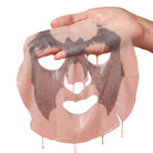 A translucent pink sheet mask with a dark charcoal-gray adhesive layer applied, being held by a hand against a plain white background.