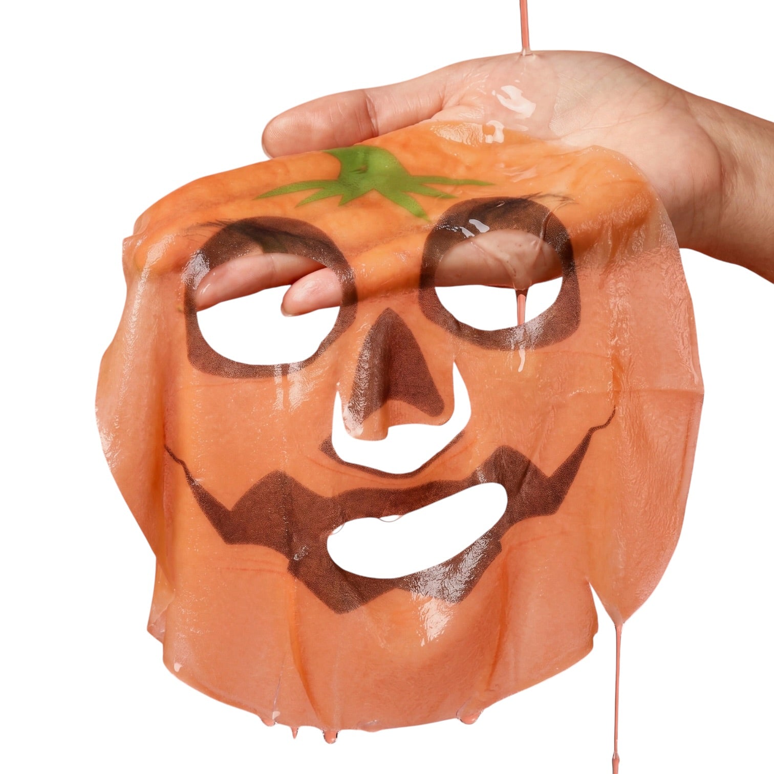 A hand holding a translucent orange sheet mask shaped like a jack-o'-lantern face with a green stem detail, shown against a plain white background.