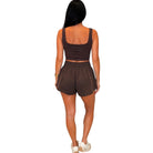 A woman shown from the back wearing a dark brown two-piece activewear set consisting of a cropped tank top and matching shorts, paired with white sneakers against a plain white background.