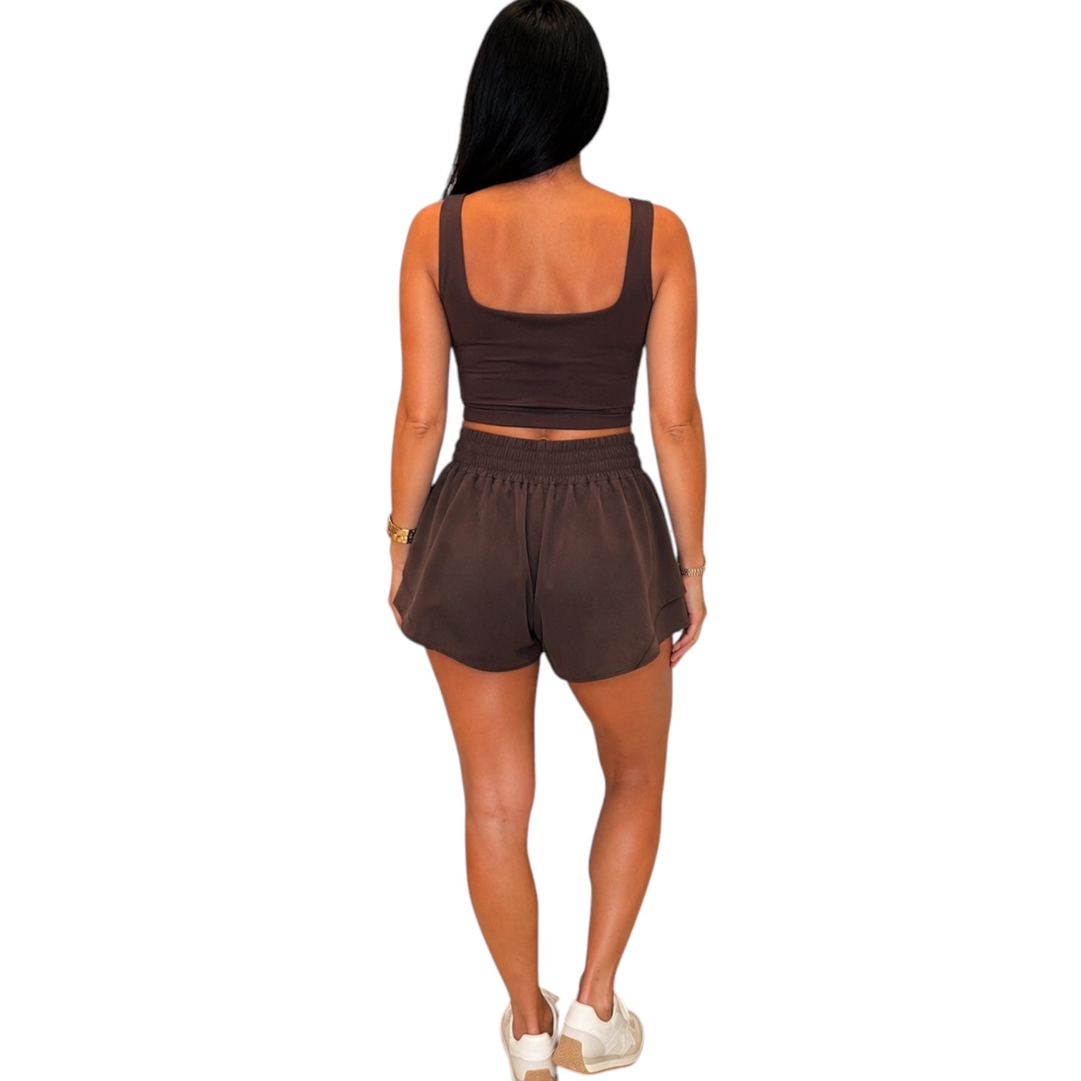 A woman shown from the back wearing a dark brown two-piece activewear set consisting of a cropped tank top and matching shorts, paired with white sneakers against a plain white background.