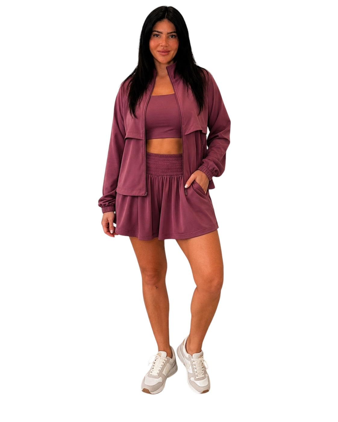 A woman modeling a matching mauve workout set consisting of a cropped tank top, high-waisted shorts, and a zip-up jacket, paired with white sneakers, against a plain white background.