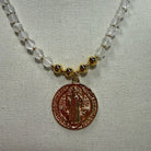 A beaded necklace featuring translucent white beads, small gold spacer beads, and a round bronze-toned religious medallion pendant, displayed on a textured white fabric background.