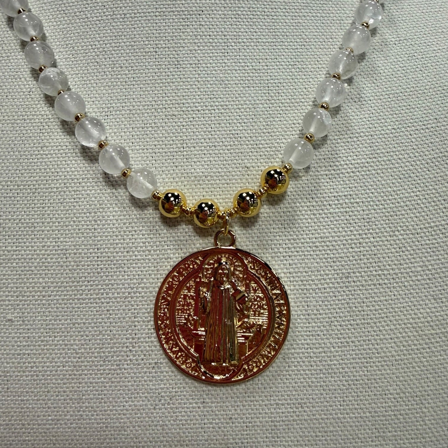 A beaded necklace featuring translucent white beads, small gold spacer beads, and a round bronze-toned religious medallion pendant, displayed on a textured white fabric background.