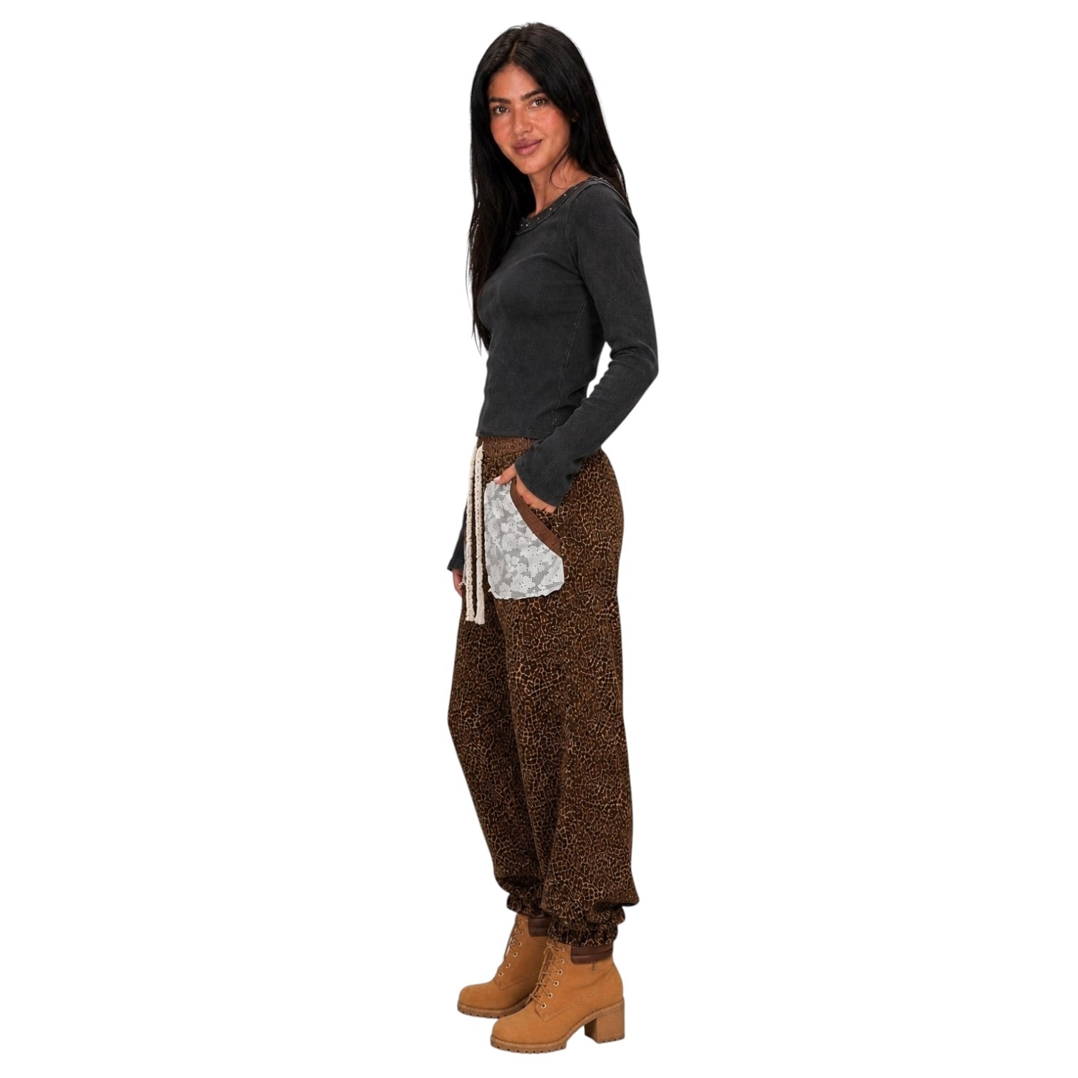 A woman modeling a pair of brown leopard-print harem pants with a white patterned pocket, paired with a dark gray long-sleeve top and tan ankle boots, against a solid white background.