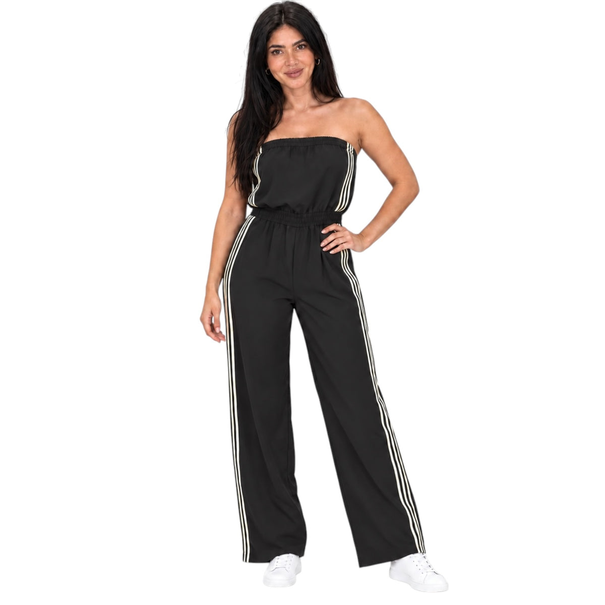 A woman modeling a black strapless jumpsuit with white side stripes, paired with white sneakers, against a plain white background.