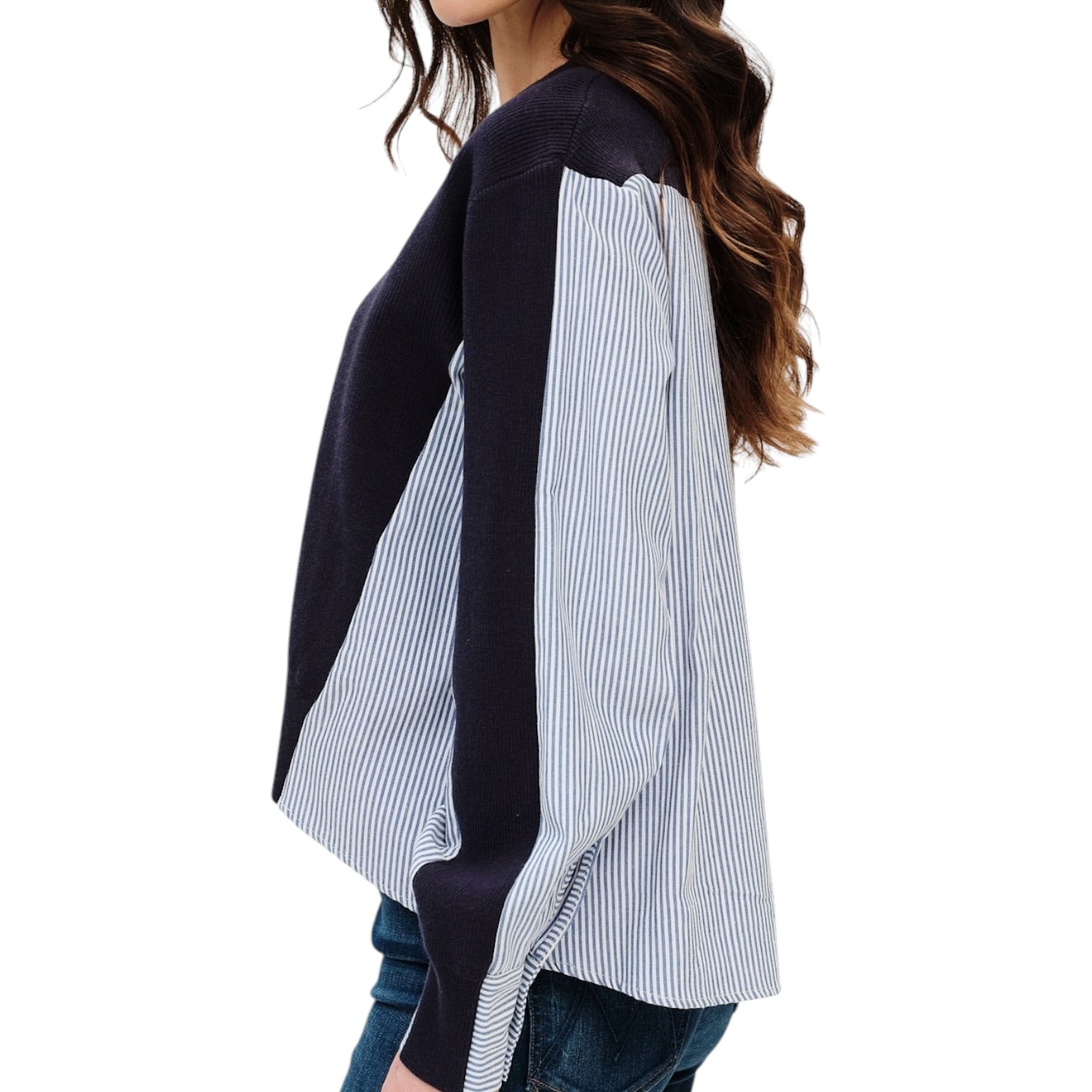 A woman wearing a color-block knit top with solid navy blue sections and light blue and white vertical stripes on the sleeves and side panels, paired with blue jeans against a plain white background.