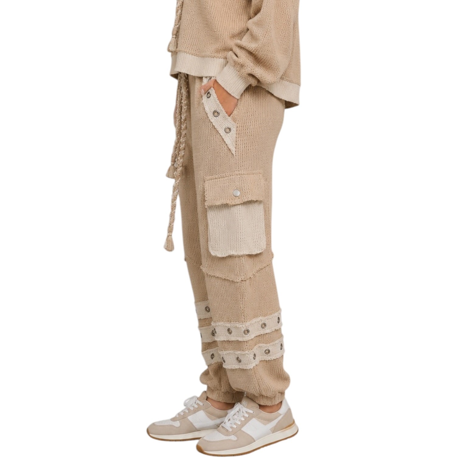 A person shown from the waist down wearing beige knit cargo jogger pants with cream fabric accents, drawstring details, and frayed trim, paired with light beige sneakers on a white background.