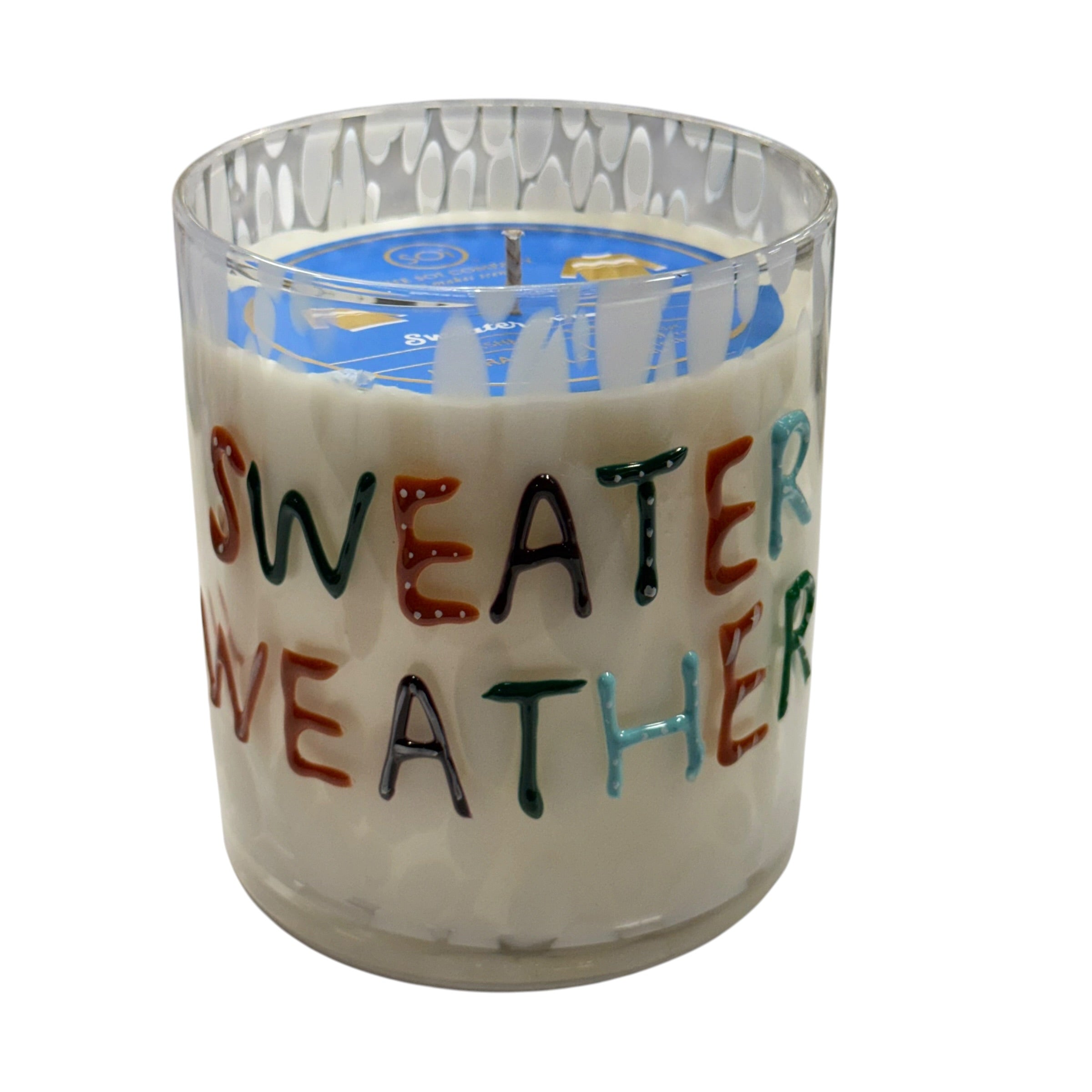 A clear glass jar candle filled with off-white wax, featuring the phrase 'SWEATER WEATHER' in multicolored hand-painted letters on the front. The candle is shown on a plain white background.