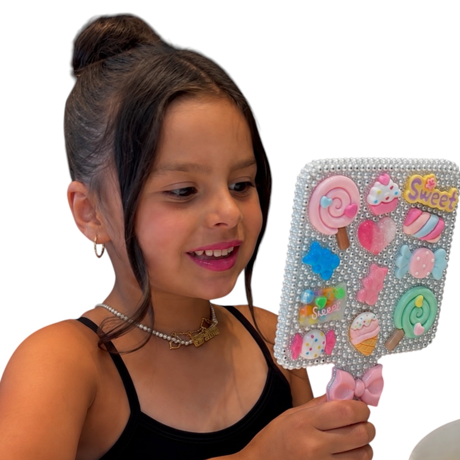 A young girl with her hair in a bun holds a decorative handheld mirror. The mirror has a rhinestone-embellished silver frame and a pink bow handle, reflecting colorful cartoon lollipops and sweets.