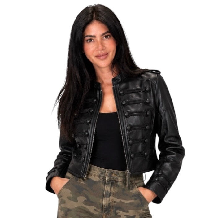 A woman modeling a black faux leather cropped jacket with military-style button details, worn over a black top and camouflage pants, against a plain white background.