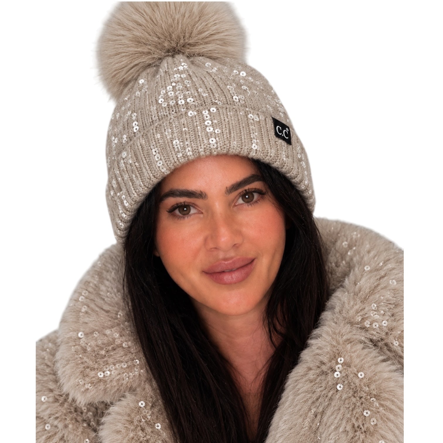 A woman wearing a beige knit beanie with silver sequins, a large beige faux-fur pom-pom, and a small black 'CC' logo patch. She is also wearing a matching beige faux-fur coat with sequins, set against a plain white background.