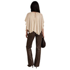 A woman shown from the back wearing a light beige draped cape-style top, dark brown flared pants, and holding a dark brown handbag against a plain white background.