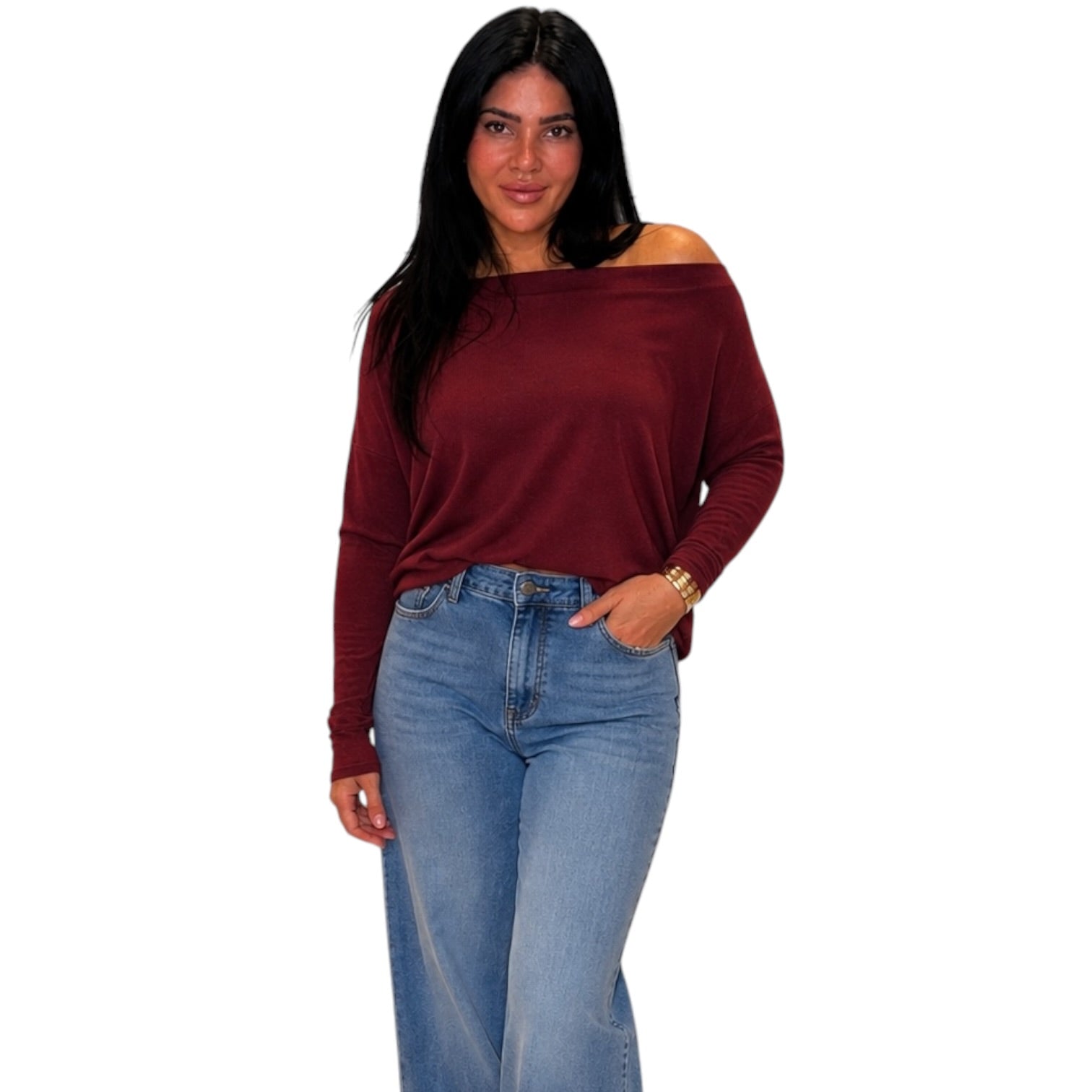 A woman modeling a long-sleeve off-the-shoulder top in burgundy, paired with light-wash blue jeans, against a plain white background.