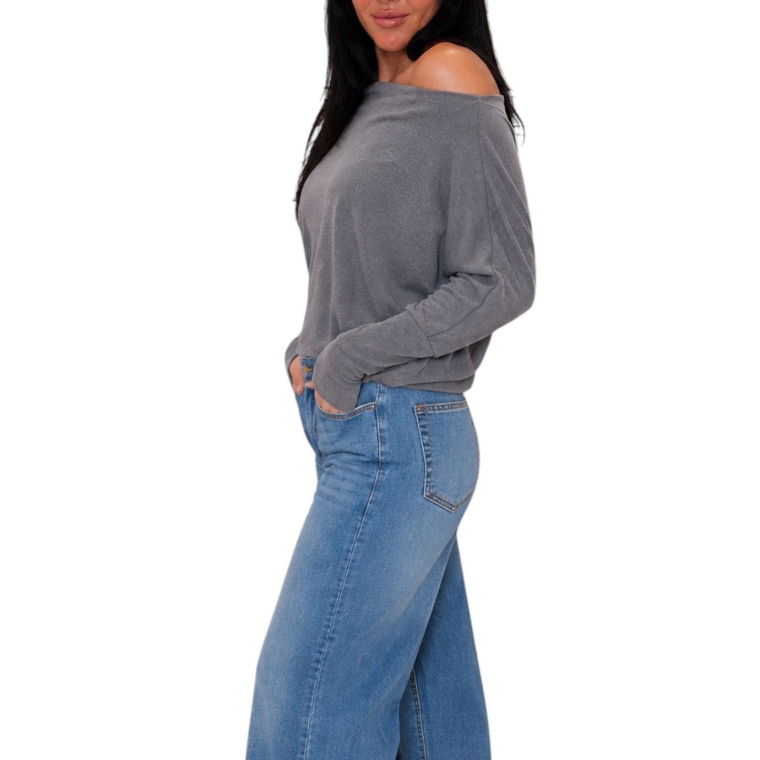 A woman wearing a gray off-the-shoulder long-sleeve knit top and blue jeans, shown from the side against a plain white background.
