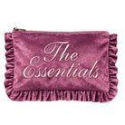 A dusty rose velvet makeup pouch with 'The Essentials' embroidered in white script across the front, featuring ruffled trim along the edges and a top zipper.