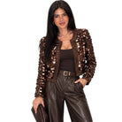 A woman modeling a dark brown sequined jacket with wide-leg brown faux-leather pants, a matching brown top, and holding a brown clutch, set against a plain white background.