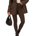 A model wearing a brown plaid blazer, dark brown leggings, and black ankle boots, holding a dark brown woven handbag against a plain white background.