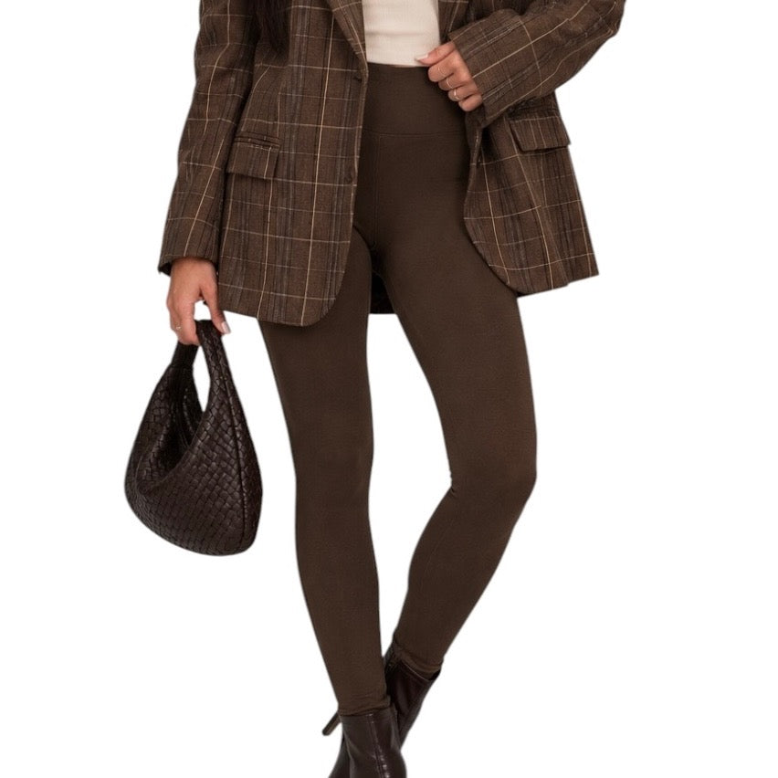 A model wearing a brown plaid blazer, dark brown leggings, and black ankle boots, holding a dark brown woven handbag against a plain white background.