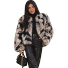 A woman modeling a black and white mottled faux fur jacket, styled with a black turtleneck, black leather pants, and a black handbag with a gold chain strap, set against a plain white background.