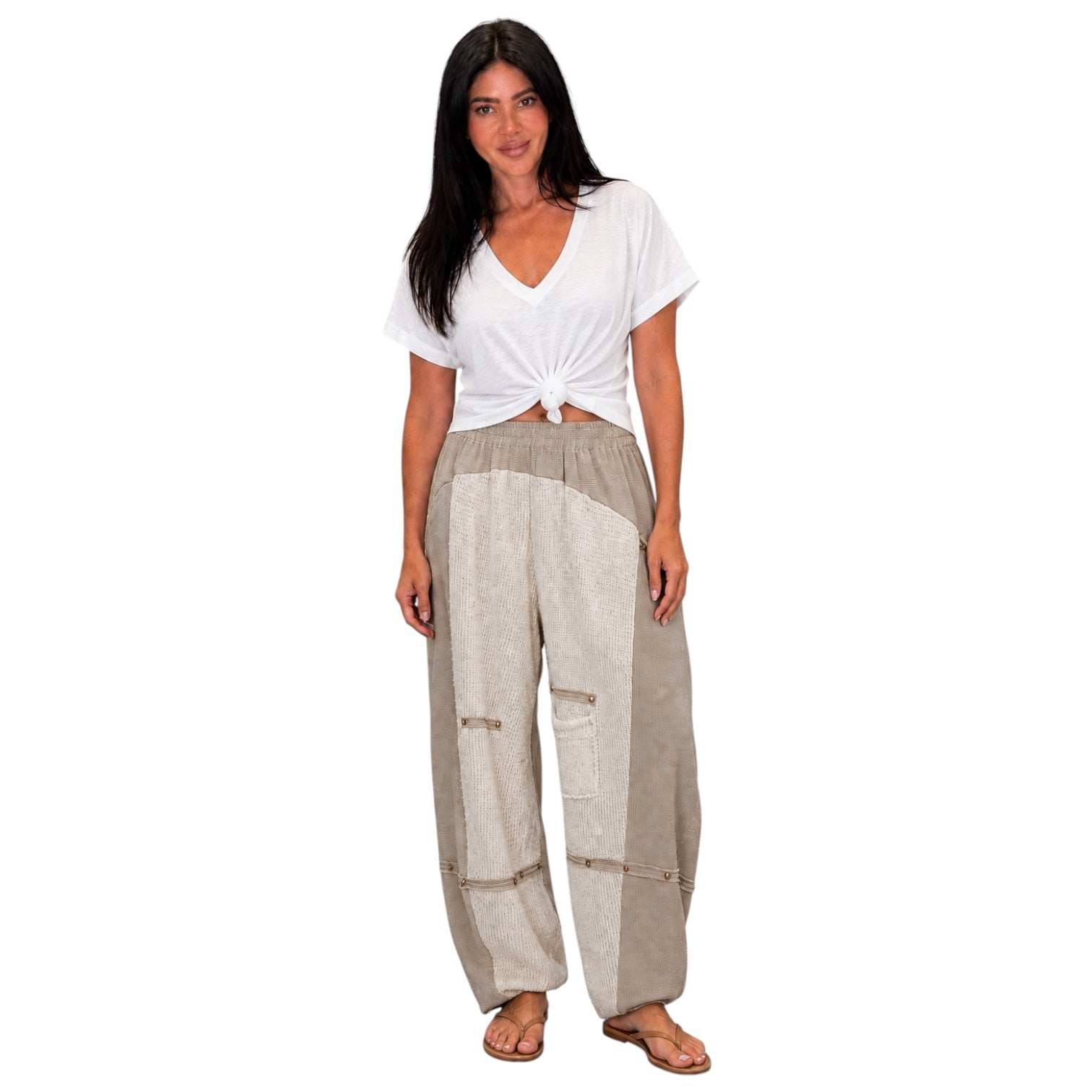 A woman modeling loose, wide-leg pants with a mixed beige and light gray linen-like texture, paired with a white tied t-shirt and brown sandals, on a solid white background.