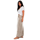 A woman modeling a pair of wide-leg beige pants with textured side panels and a white tied t-shirt, standing against a solid white background.
