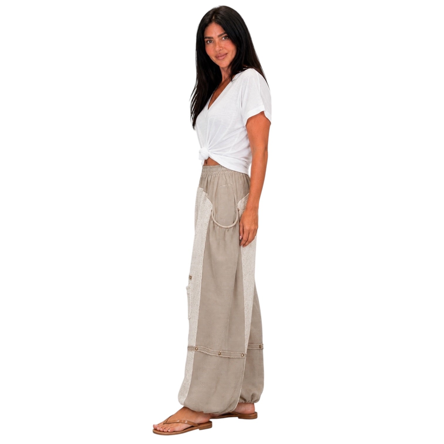A woman modeling a pair of wide-leg beige pants with textured side panels and a white tied t-shirt, standing against a solid white background.