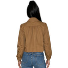 A woman viewed from the back wearing a cropped tan jacket with gathered detailing at the shoulders and blue jeans, isolated on a white background.