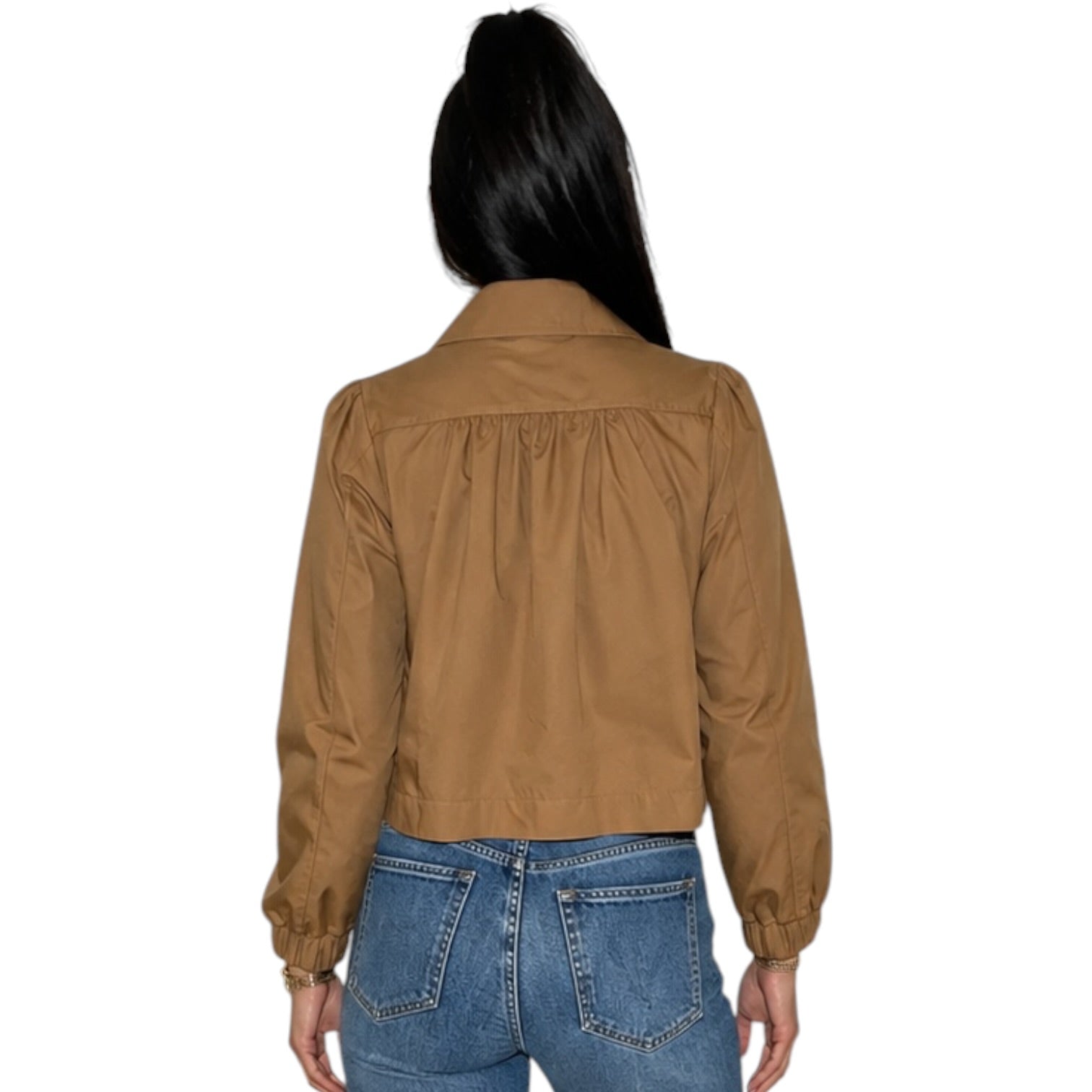A woman viewed from the back wearing a cropped tan jacket with gathered detailing at the shoulders and blue jeans, isolated on a white background.