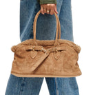 A tan suede handbag with a fluffy shearling-style lining and double top handles, held by a person wearing blue jeans against a plain white background.