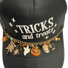 A black trucker hat with 'TRICKS and treats' printed in white on the front, decorated with a gold chain across the brim featuring various Halloween charms including a spider, ghost, pumpkin, and sugar skull.