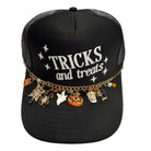 A black trucker hat with 'TRICKS and treats' printed on the front, decorated with a gold chain and various Halloween-themed charms including a pumpkin, ghost, skull, spider, and candy.