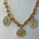 Gold chain necklace with three oval pendants featuring gold Miraculous Medal motifs set in white shell-like centers and bordered with small clear stones, displayed on a textured off-white fabric background.